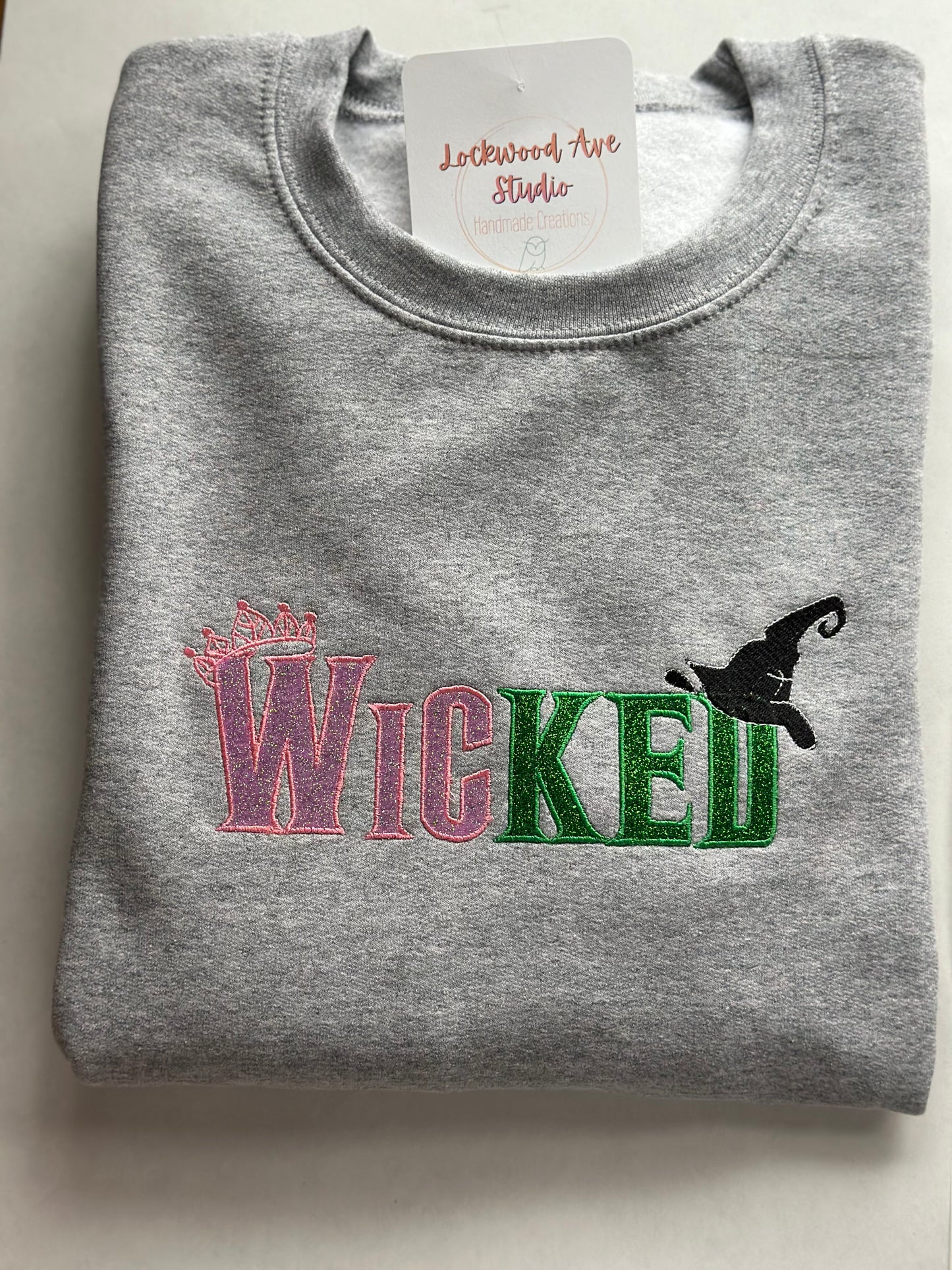 Gray sweatshirt with pink and green embroidered “WICKED” text, featuring a small crown above the W and a black witch hat above the D, with a Lockwood Ave Studio tag tucked into the collar.