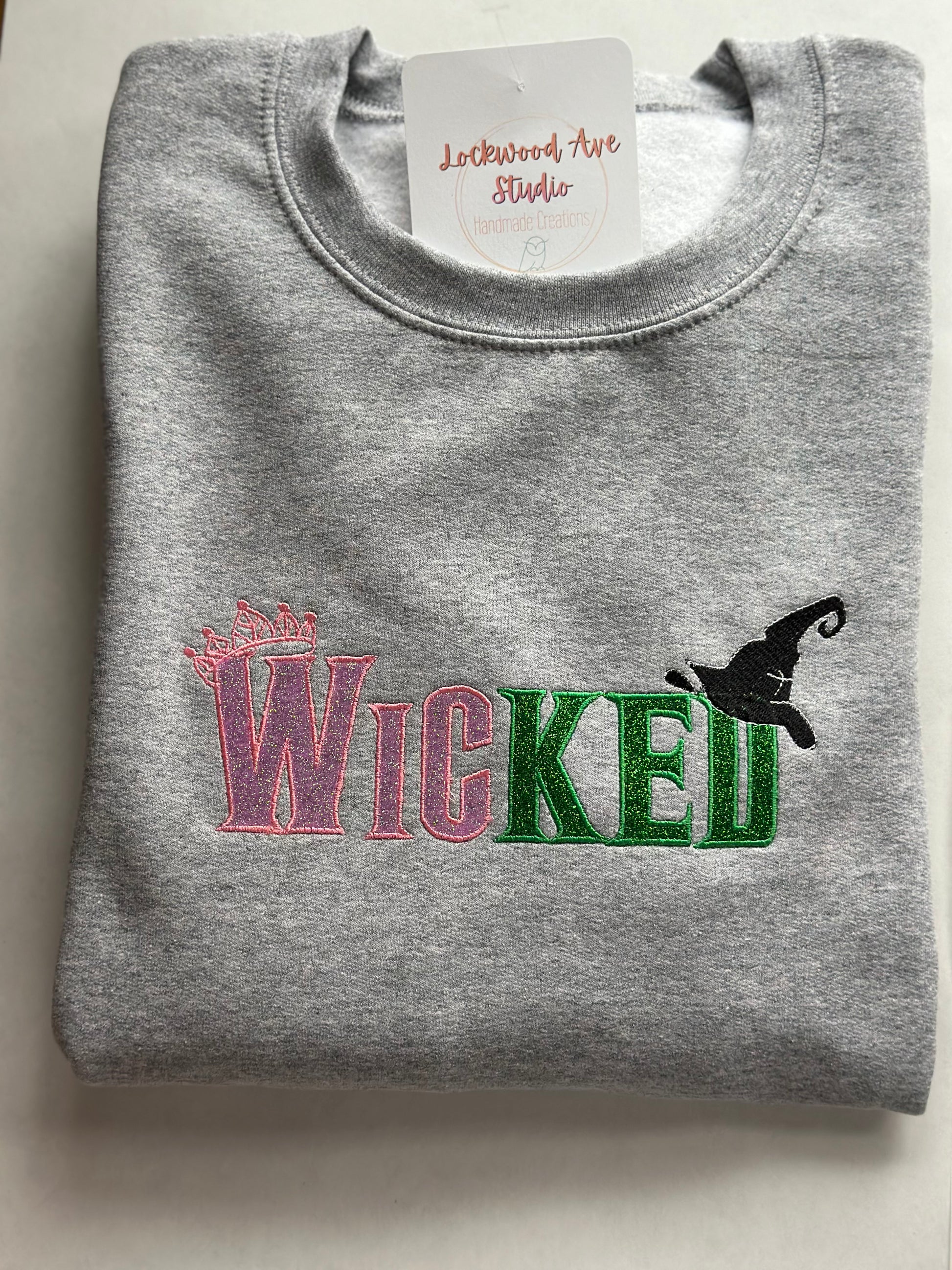 Gray sweatshirt with pink and green embroidered “WICKED” text, featuring a small crown above the W and a black witch hat above the D, with a Lockwood Ave Studio tag tucked into the collar.