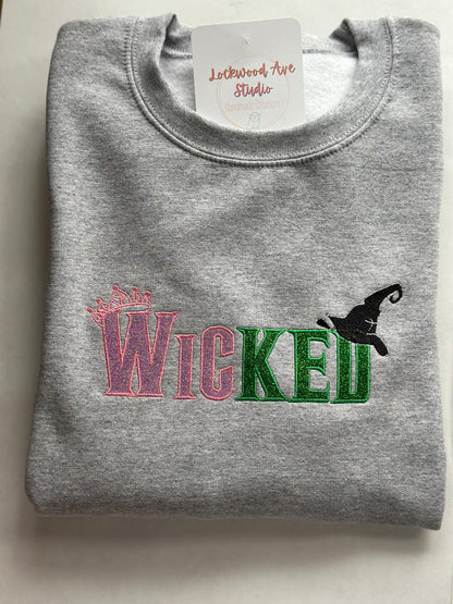 Gray sweatshirt with pink and green embroidered “WICKED” text, featuring a small crown above the W and a black witch hat above the D, with a Lockwood Ave Studio tag tucked into the collar.