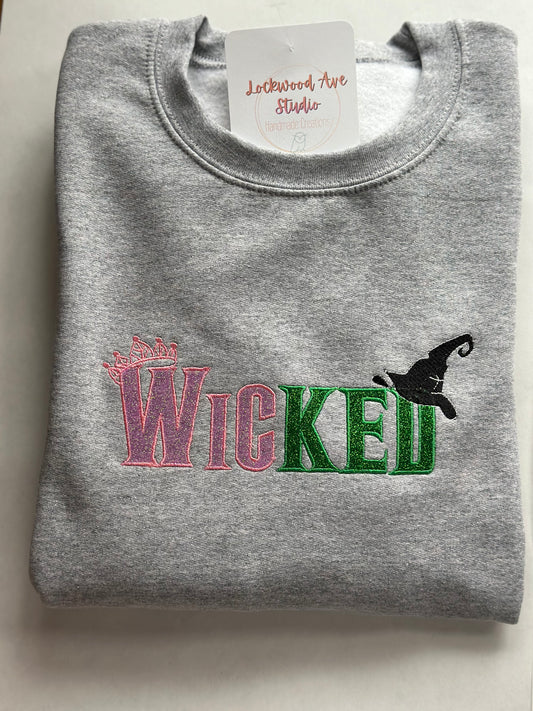 Gray sweatshirt with pink and green embroidered “WICKED” text, featuring a small crown above the W and a black witch hat above the D, with a Lockwood Ave Studio tag tucked into the collar.