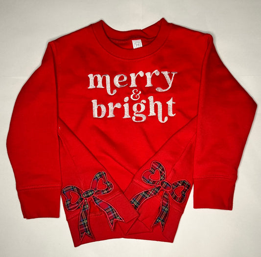 Red children’s sweatshirt with white “merry & bright” embroidery on the chest and two plaid side bow appliqués with heart-shaped details near the hem.
