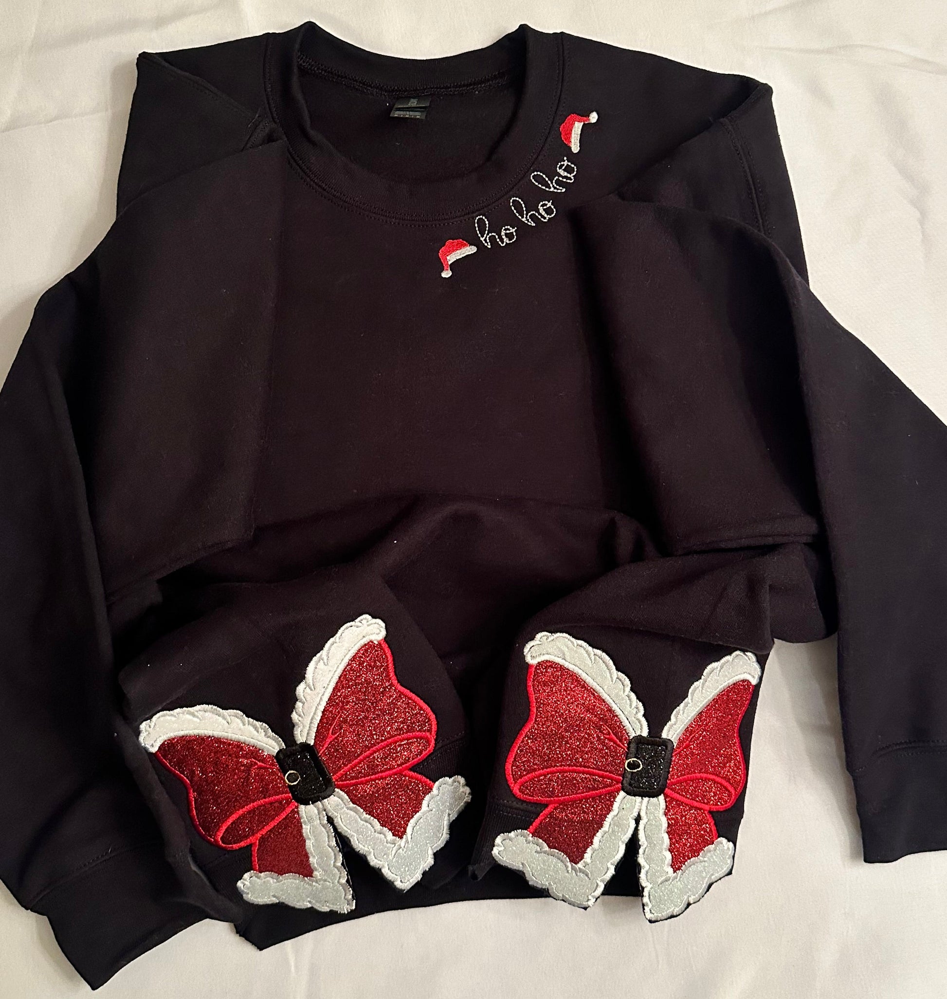 Children’s black sweatshirt featuring “ho ho ho” embroidery with Santa hats on the neckline and two glittery red Santa bow appliqués with white trim and buckle accents on the sides.