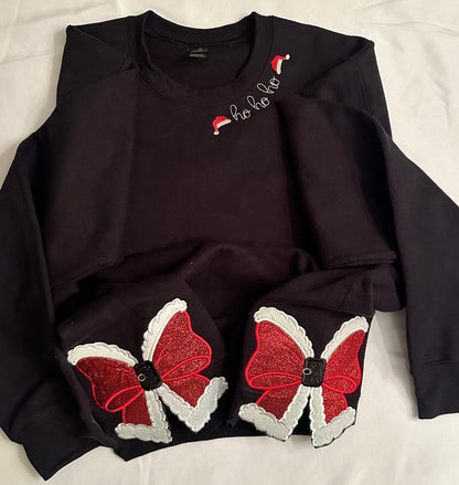 Children’s black sweatshirt featuring “ho ho ho” embroidery with Santa hats on the neckline and two glittery red Santa bow appliqués with white trim and buckle accents on the sides.
