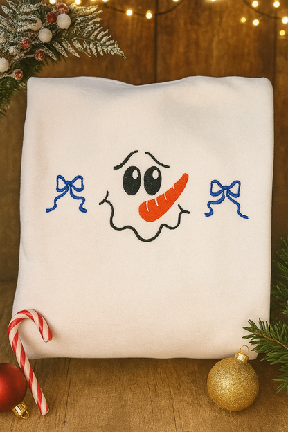 Folded white sweatshirt with embroidered snowman face featuring a carrot nose, black eyes, and two blue bows, surrounded by festive Christmas lights, candy cane, and ornaments.