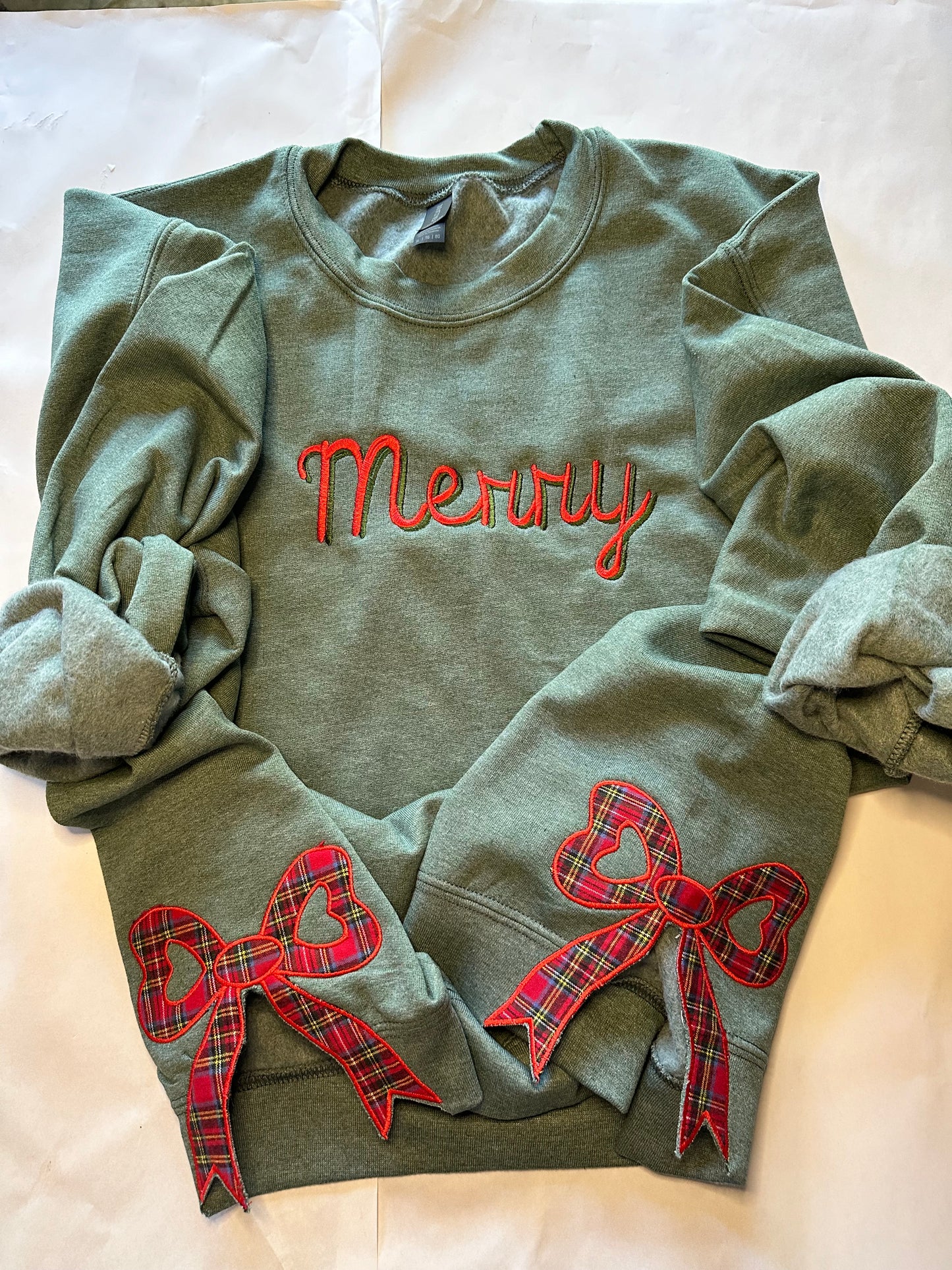 Green sweatshirt with “Merry” embroidered in red  and green across the chest and two red plaid side bow appliqués with heart-shaped centers, displayed on a light background.
