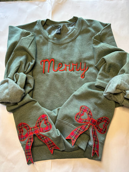 Green sweatshirt with “Merry” embroidered in red  and green across the chest and two red plaid side bow appliqués with heart-shaped centers, displayed on a light background.