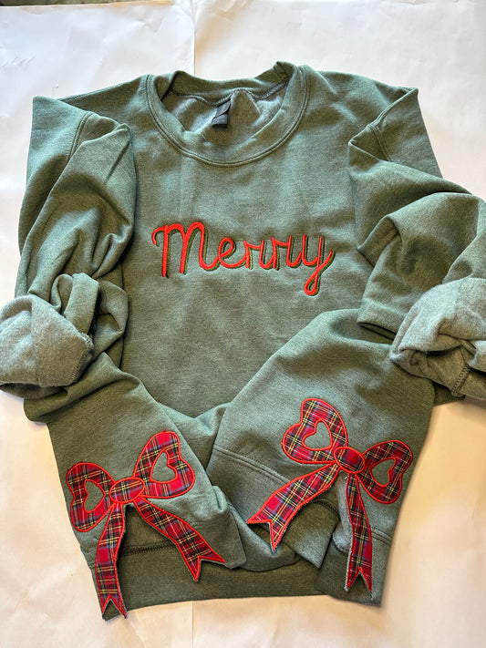 Green sweatshirt with “Merry” embroidered in red  and green across the chest and two red plaid side bow appliqués with heart-shaped centers, displayed on a light background.