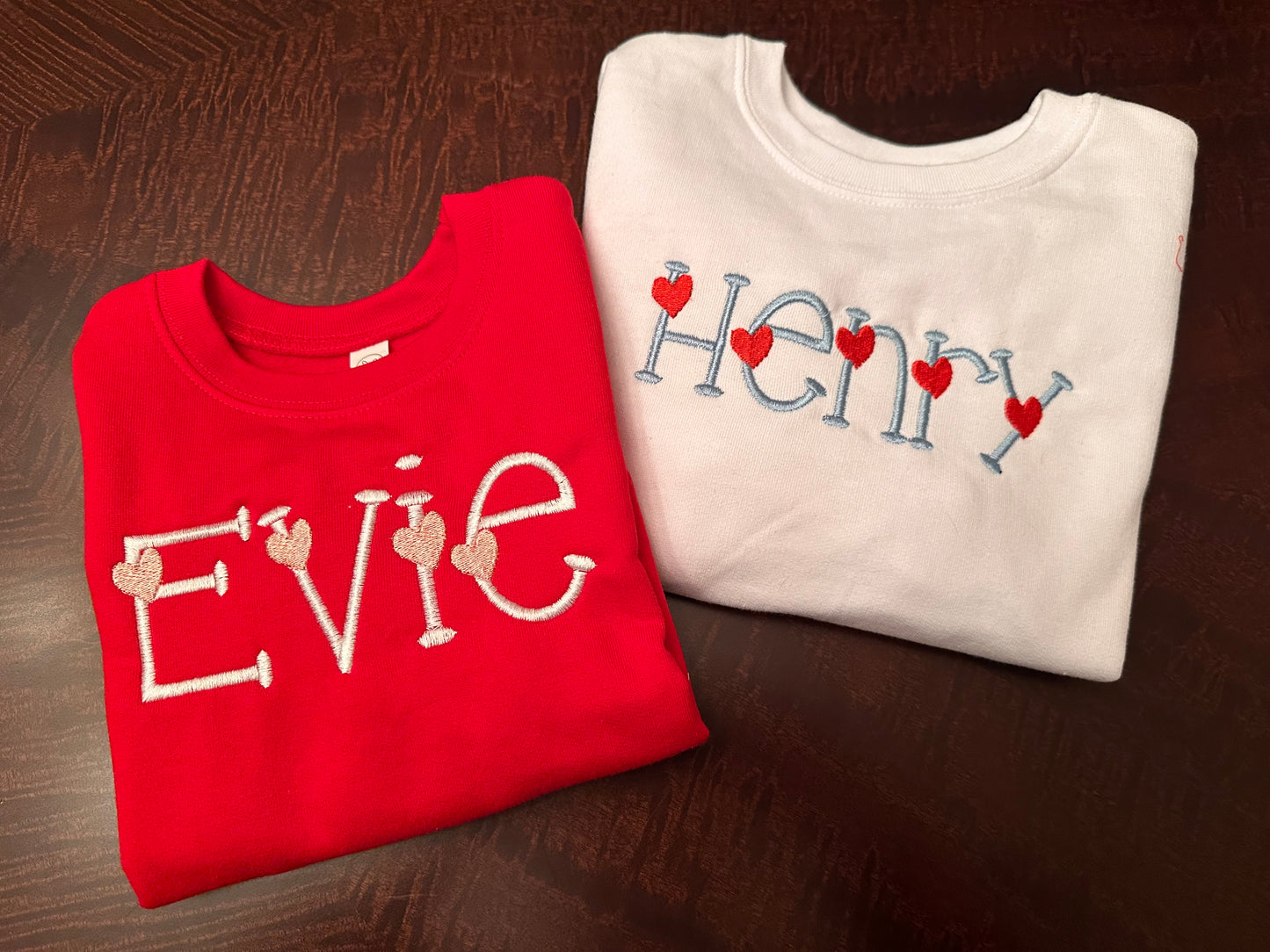 Personalized Valentine’s Day embroidered sweatshirts for kids featuring custom names with heart-themed designs, showing Evie in white lettering with light pink hearts on a red sweatshirt.  Henry in light blue with red hearts on a white sweatshirt.  