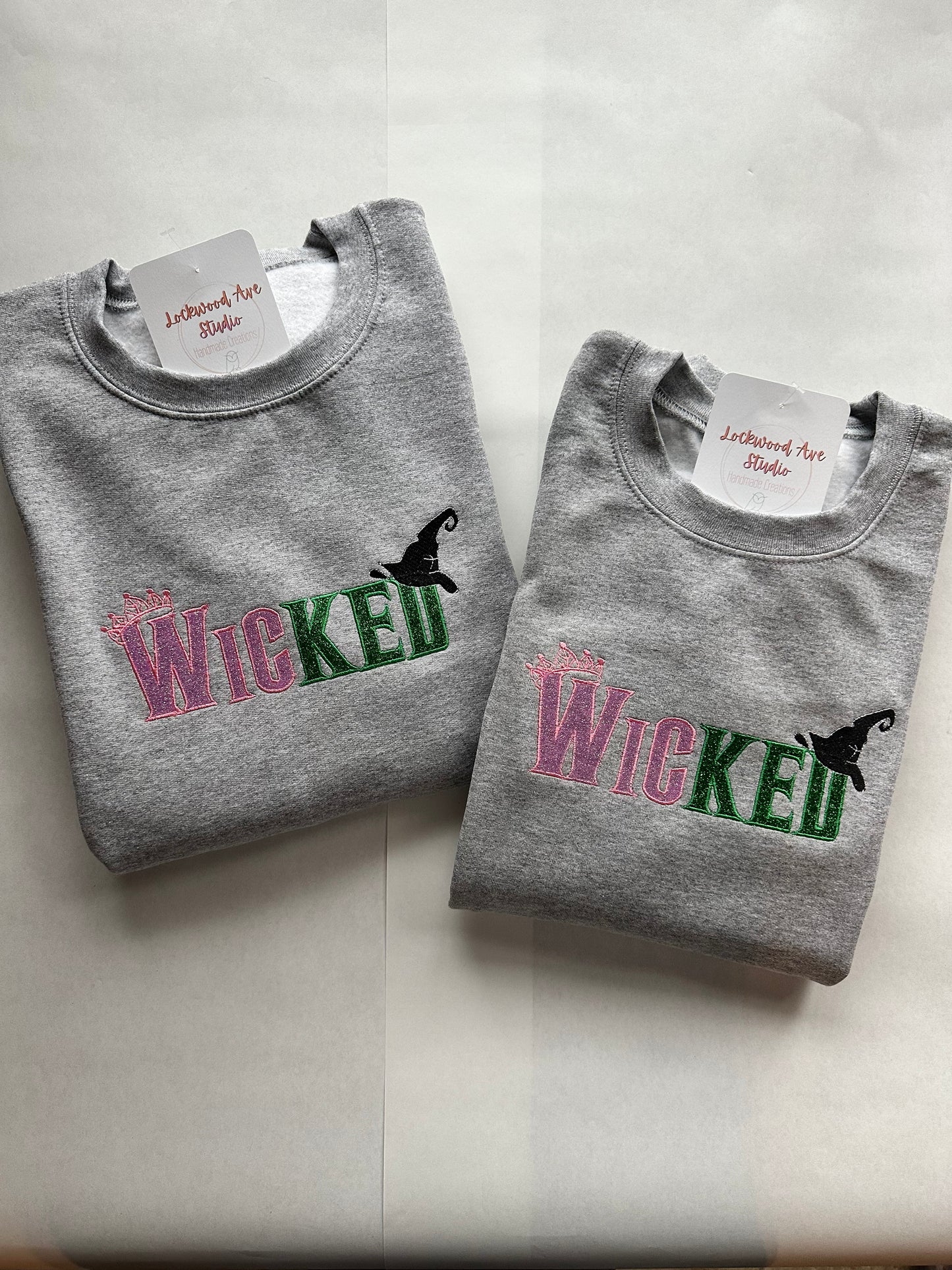 Two Gray sweatshirts  with pink and green embroidered “WICKED” text, featuring a small crown above the W and a black witch hat above the D, with a Lockwood Ave Studio tag tucked into the collar.